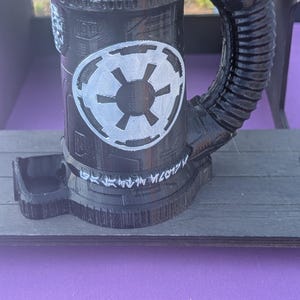 May include: Black 3D-printed mug with a cylindrical body and a large handle. The mug features a white Imperial symbol and what appears to be Aurebesh text. The mug is sitting on a wooden surface.
