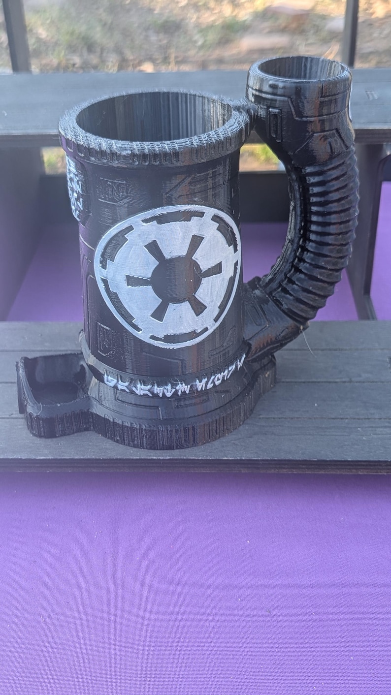 May include: Black 3D-printed mug with a cylindrical body and a large handle. The mug features a white Imperial symbol and what appears to be Aurebesh text. The mug is sitting on a wooden surface.