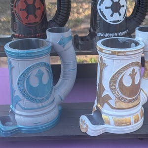 May include: Collection of detailed, themed mugs. The mugs feature designs with the Imperial and Rebel symbols in black, red, white, and gold. The mugs have a cylindrical shape with a handle and a base.
