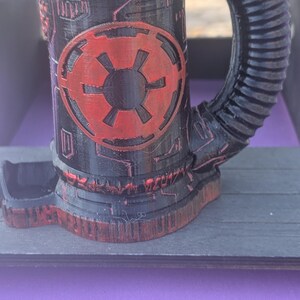 May include: Black and red 3D-printed mug with a unique design. The mug features a cylindrical body with a handle and a base. A red emblem is prominently displayed on the front. The design has a futuristic, mechanical aesthetic.