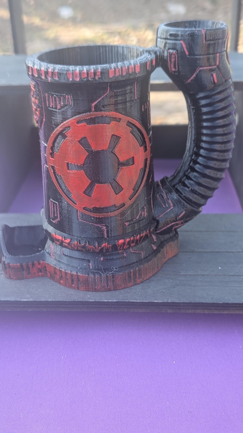 May include: Black and red 3D-printed mug with a unique design. The mug features a cylindrical body with a handle and a base. A red emblem is prominently displayed on the front. The design has a futuristic, mechanical aesthetic.
