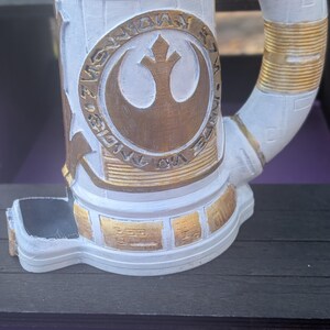 May include: White and gold-painted stein featuring the Rebel Alliance symbol. The mug has a cylindrical body with a curved handle and decorative gold accents. The design includes a detailed emblem and text.