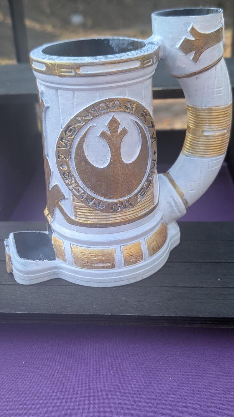 May include: White and gold-painted stein featuring the Rebel Alliance symbol. The mug has a cylindrical body with a curved handle and decorative gold accents. The design includes a detailed emblem and text.