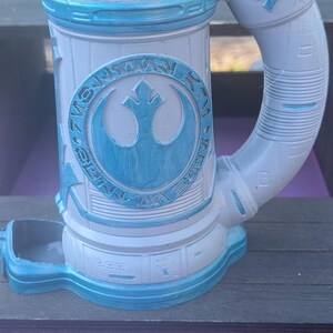 May include: A white and blue ceramic mug with a handle, featuring a circular emblem with a bird-like symbol. The mug has a cylindrical shape with a wide opening and a base. The design includes blue accents and text.
