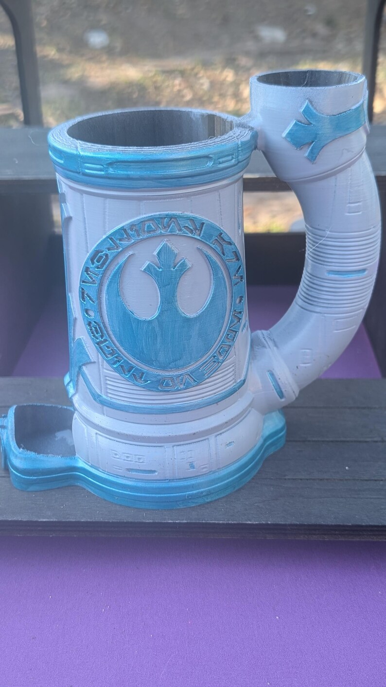 May include: A white and blue ceramic mug with a handle, featuring a circular emblem with a bird-like symbol. The mug has a cylindrical shape with a wide opening and a base. The design includes blue accents and text.