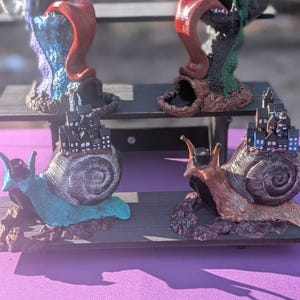 May include: A display of whimsical fantasy figurines. Two monster-like creatures with open mouths and long tongues, and two snail figurines with miniature cityscapes on their shells. The figurines are on a black shelf against a purple background.