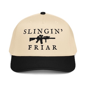 Puede incluir: Gorra de béisbol con corona color crema y visera negra. La gorra presenta el texto "SLINGIN' FRIAR" encima y debajo de una silueta negra de un rifle.