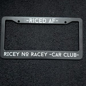 May include: Black license plate frame with white text that reads "-RICED AF-" at the top and "RICEY NO RACEY -CAR CLUB-" at the bottom. The frame has a rectangular shape with rounded corners and mounting holes.