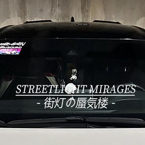 May include: A black car windshield with a white decal that reads "STREETLIGHT MIRAGES" in English and Japanese. A small Pikachu plush and a spider decoration are visible.