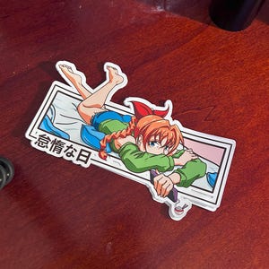 May include: A sticker featuring an anime-style illustration of a girl with orange hair, wearing a green top and blue shorts, lying on a bed. The sticker has Japanese text and a white border. The background is a mix of blue and pink.