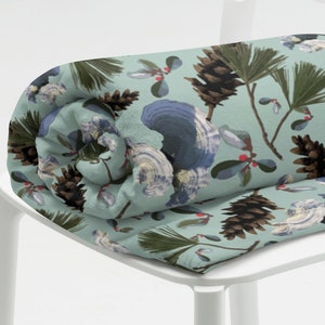 Könnte beinhalten: Ein zusammengerolltes blaues Dekokissen mit einem Muster aus Tannenzapfen, Tannenzweigen und roten Beeren. Das Kissen liegt auf einem weißen Stuhl.