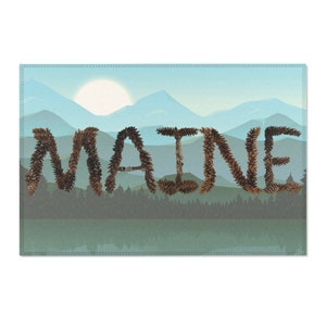 Könnte beinhalten: Ein rechteckiger dekorativer Teppich mit dem Wort "MAINE", das mit braunen Tannenzapfen geschrieben wurde. Der Hintergrund zeigt eine Landschaft mit Bergen, einem See und einer aufgehenden Sonne in Blau- und Grüntönen.