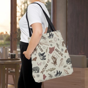 May include: A beige tote bag with a black strap features a repeating pattern of illustrations of moose, bears, fish, and the state of Maine. The text "Maine" is repeated on the bag.