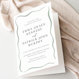 May include: A white wedding invitation with a sage green wavy border. The text reads "Emma Grace Kennedy and Patrick John Murphy" with the wedding date and location. The invitation is folded and set against a neutral fabric.