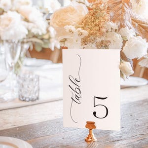 May include: White card with black calligraphy that says "Table 5" on a gold metal stand. The card is sitting on a wooden table with white flowers in the background.