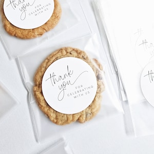 May include: Two clear plastic bags containing cookies. Each cookie has a white round sticker with black text that reads "Thank you for celebrating with us".
