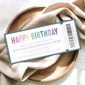 May include: A birthday gift voucher with the words "HAPPY BIRTHDAY" in rainbow colors. The voucher includes space for a gift or destination, and additional information. The voucher is on a wooden plate with a beige cloth.