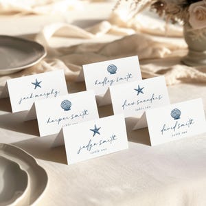 May include: White place cards with navy blue seashell and starfish illustrations. Each card features a name in elegant script. Some cards also include table numbers. The cards are arranged on a cream-colored tablecloth.