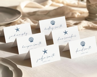 Può includere: Segnaposto bianchi con illustrazioni di conchiglie e stelle marine blu navy. Ogni biglietto presenta un nome in elegante corsivo. Alcuni biglietti includono anche i numeri dei tavoli. I biglietti sono disposti su una tovaglia color crema.
