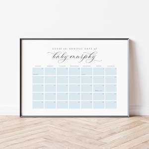 May include: A framed calendar print with a white background and a black border. The calendar has light blue squares for each day and the text "Guess the Arrival Date of baby murphy" in elegant script. The calendar is a baby shower game.