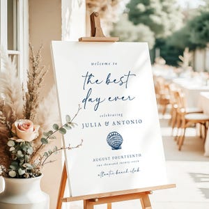 Puede incluir: Un letrero de boda blanco sobre un caballete de madera. El texto en azul marino dice "welcome to the best day ever", los nombres Julia & Antonio, un gráfico de concha y la fecha Catorce de agosto.