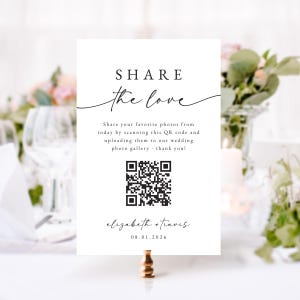 May include: A white wedding sign with the text "SHARE the love" in elegant script. It includes a QR code for photo sharing, the names "elizabeth + travis", and the date 08.01.2026. The sign is held by a gold-colored clip.