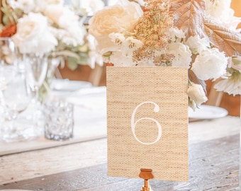 Könnte beinhalten: Eine Tischkarten-Nummer sechs mit gewebter Textur, in einem goldfarbenen Halter. Die Karte ist beige mit einer großen weißen Zahl sechs. Der Hintergrund umfasst ein Blumenarrangement und eine Tischdekoration.