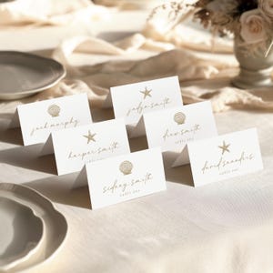 May include: White place cards with gold seashell and starfish designs. Each card displays a name in elegant script, with the words "table one" below. The cards are arranged on a cream-colored tablecloth, suggesting a beach or nautical theme.