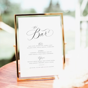 May include: A gold framed sign with a white background and black text that reads "the Bar" with a list of beer, wine, and liquor options. The sign is on a wooden surface.