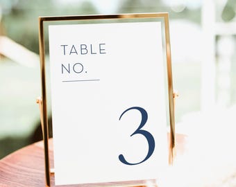 May include: A gold-framed table number sign with the text "TABLE NO." and the number "3" in navy blue. The sign is displayed on a wooden surface, suitable for weddings or events.