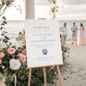 Könnte beinhalten: Ein weißes Hochzeits-Willkommensschild auf einer Holzstaffelei. Das Schild hat marineblauen Text mit der Aufschrift "Willkommen zur Hochzeit von Julia Grace und Antonio James, vierzehnter August, zweitausendsiebenundzwanzig". Eine Muschelgrafik ist darunter.