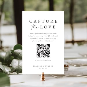 May include: A white wedding sign with the text "CAPTURE the LOVE" in a stylish font. It includes a QR code and instructions to share photos. The sign is held by a gold-coloured clip and features the names Isabella & Alex and the date 09.05.26.