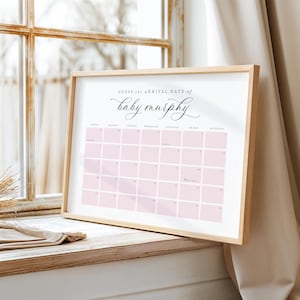 May include: A framed calendar print with a light wood frame. The calendar has a pink grid with the text "Guess the Arrival Date of baby murphy" in elegant script. The calendar is displayed on a wooden surface near a window.
