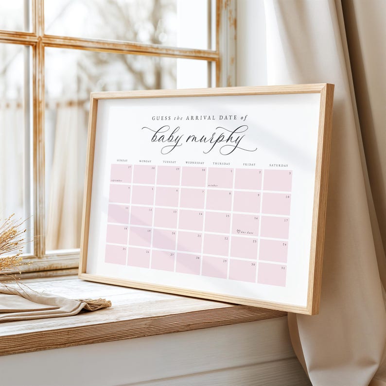 May include: A framed calendar print with a light wood frame. The calendar has a pink grid with the text "Guess the Arrival Date of baby murphy" in elegant script. The calendar is displayed on a wooden surface near a window.