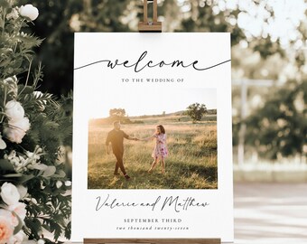 May include: A white wedding welcome sign on a wooden easel. The sign features the word "welcome" in elegant script, with the text "TO THE WEDDING OF" below. A photo of a couple is centered, with names and the wedding date below.