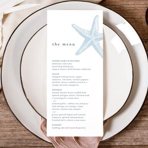 May include: A menu card featuring a light blue starfish design rests on a white plate with a gold rim. The menu details starters, salad, main courses, and desserts. The text is in a dark blue font.