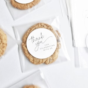 May include: A clear plastic bag containing a round, brown cookie. The cookie has a white circular sticker with the text "Thank you for celebrating with us".