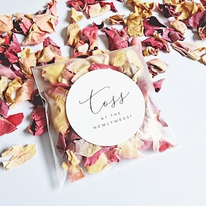 May include: A transparent bag filled with dried flower petals in red, pink, and yellow hues. A white circular label on the bag displays the text "Toss at the Newlyweds!" in black script. Loose petals are scattered around the bag.