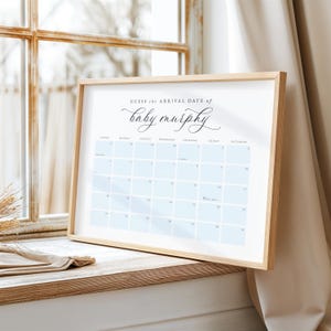 May include: A framed calendar print with the text "Guess the Arrival Date of baby murphy" in elegant script. The calendar grid features light blue squares for each day. The frame is made of light-colored wood.