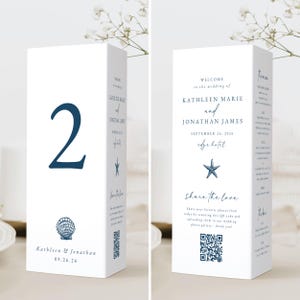 Op de afbeelding: Een witte trouwtafelnummerkaart met een groot marineblauw nummer 2. De kaart heeft een schelpgrafiek, de namen Kathleen & Jonathan en de datum 09.26.26. De andere kant zegt "Welkom bij de bruiloft van Kathleen Marie en Jonathan James."