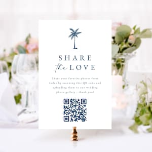 May include: A white wedding sign with navy blue text and a palm tree illustration. The sign reads "SHARE the LOVE" and includes a QR code. The sign is held by a gold-colored clip.