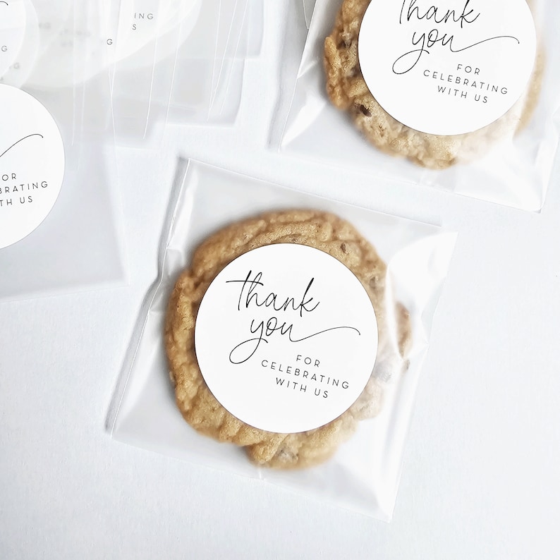 May include: A clear plastic bag containing a chocolate chip cookie. The cookie has a white round sticker on it that says "Thank you for celebrating with us".