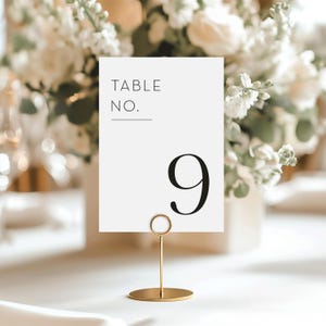 May include: A white table number card with the text "TABLE NO." and the number "9" in black. The card is held by a gold metal stand with a circular base. The background features white flowers.