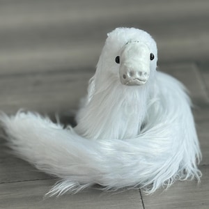 May include: A white, fluffy plush toy resembling a mythical creature, possibly a dragon or serpent. It features a detailed head with black eyes and a long, curled tail. The toy is designed to sit on a surface, with a soft, textured body.