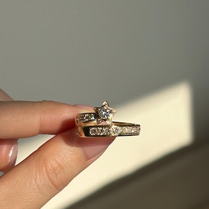 May include: A gold-colored ring set featuring a star-shaped center stone and smaller stones along the bands. The rings are held up, showcasing their design and sparkle. The jewelry is set against a neutral background.