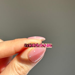 Peut inclure: Une bague en or ornée d'une rangée continue de pierres précieuses roses vives. La bague est tenue entre les doigts sur un fond gris neutre, mettant en valeur le design et la couleur de la bague.