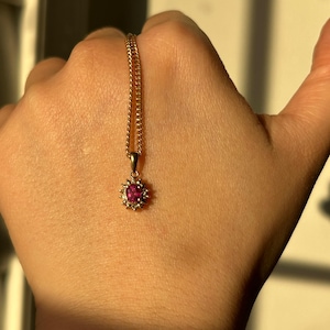 Könnte beinhalten: Eine goldfarbene Halskette mit einem Anhänger, der einen zentralen rosa Edelstein und kleinere klare Steine ​​aufweist. Der Anhänger hängt an einer filigranen Kette. Die Halskette wird vor einem hautfarbenen Hintergrund präsentiert.