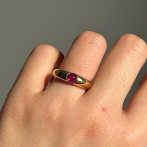 Könnte beinhalten: Ein goldener Ring mit einem Rubin-Edelstein. Der Ring hat ein glattes, abgerundetes Band und eine Zargenfassung, die den oval geformten, tiefroten Rubin hält. Der Ring wird an einer Hand gezeigt.