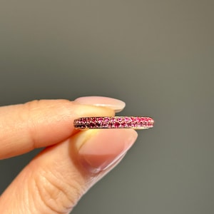 Peut inclure: Une bague en or ornée de nombreux petits pierres précieuses roses vives. La bague est tenue entre les doigts, mettant en valeur les détails délicats du bijou. Les pierres sont rapprochées, créant une ligne de couleur continue.