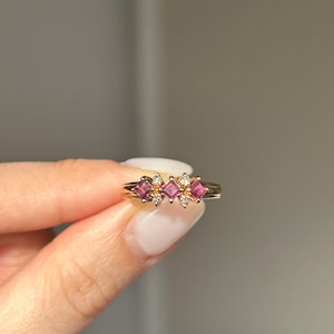 May include: A gold ring with three square, pink gemstones and small, clear accent stones. The ring is held up, displaying the design and the polished gold band. The gemstones are set in a row, creating a delicate and elegant look.